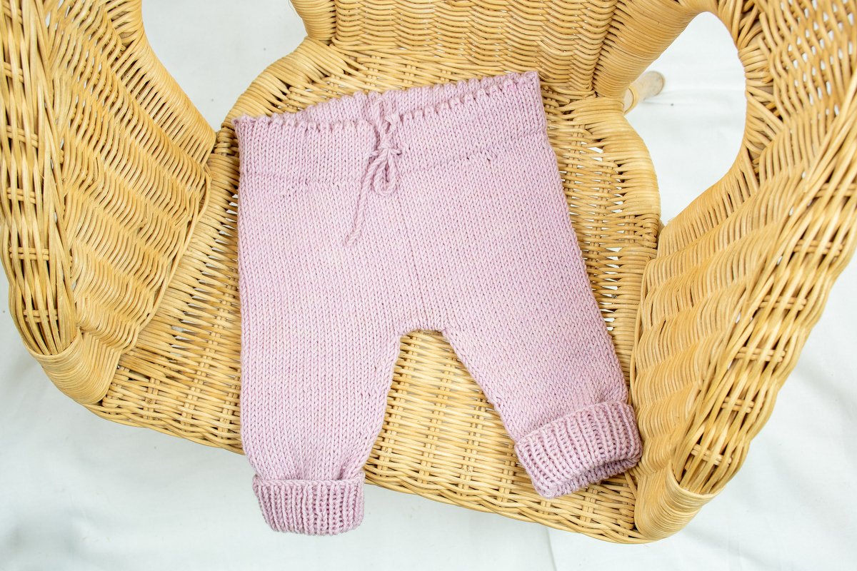 Gestrickte Babyhose in Flieder mit Kordelzug und gerippten Bündchen auf einem Korbstuhl