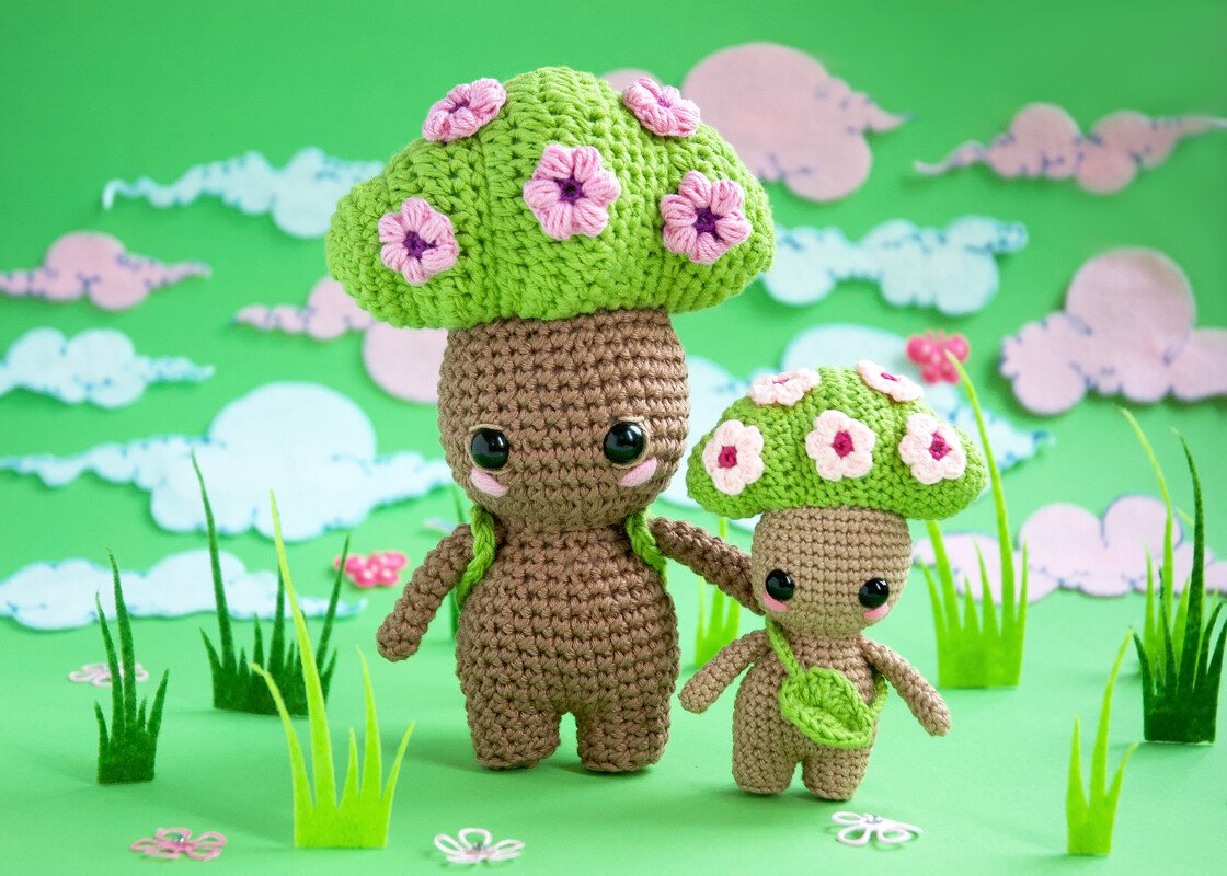 Two crocheted amigurumi mushroom dolls with green caps decorated with pink flowers on a green backdrop with felt grass.