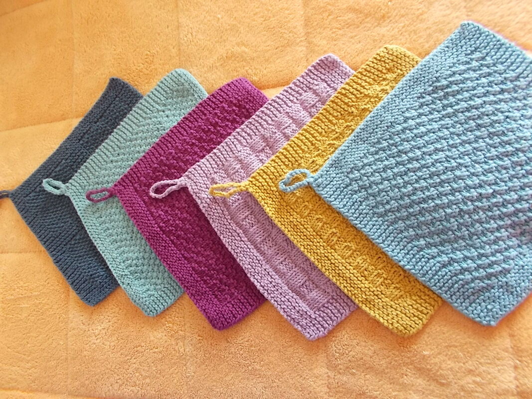 Reihe von sechs gestrickten Topflappen in verschiedenen Farben mit Aufhängeschlaufen auf orangefarbener Unterlage
