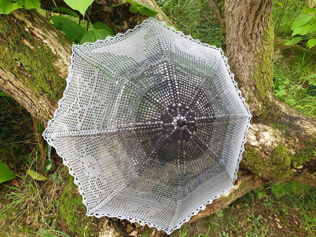 Gehäkelter Sonnenschirm in Grau-Weiß mit Spitzenmuster auf moosigem Baumstamm