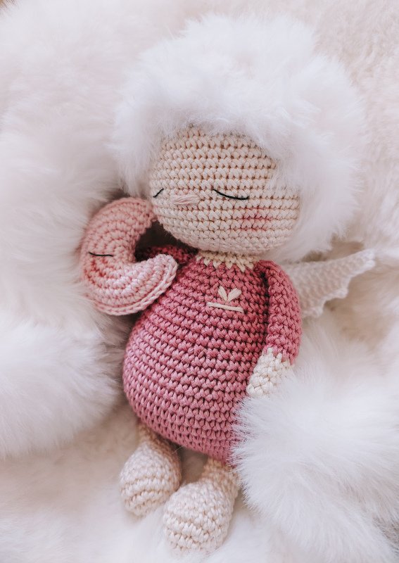 Sleeping crocheted amigurumi angel doll in a pink dress with fluffy white hair and small wings holding a crescent moon.