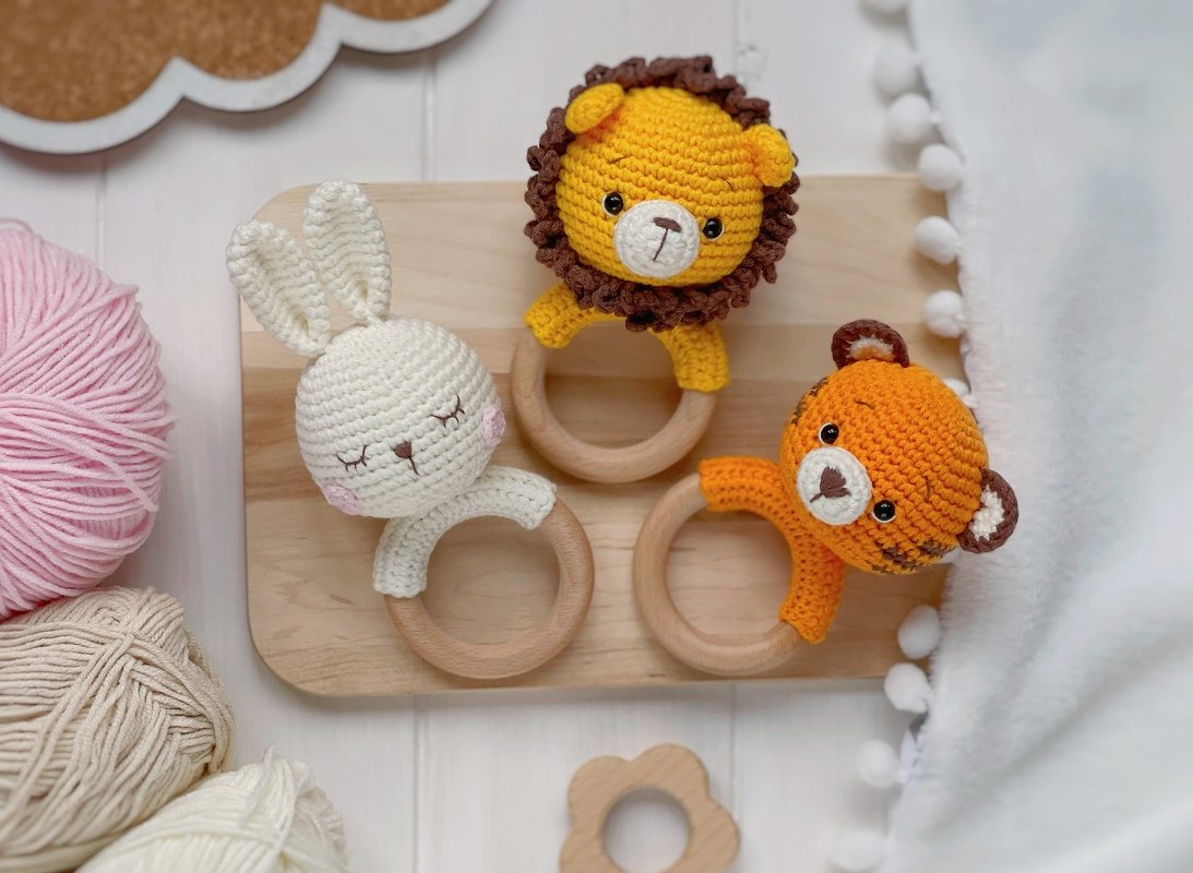 Three crocheted animal baby rattles with wooden rings: white bunny, yellow lion, and orange tiger on a wooden board with yarn and a blanket.