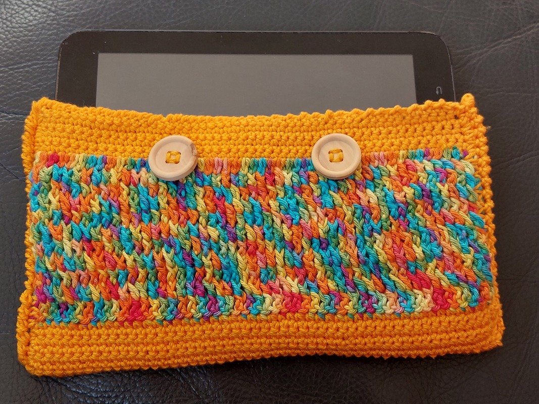 Gehäkelte orangefarbene Tablet-Hülle mit buntem Mittelstreifen und zwei hellen Holzknöpfen; Tablet teilweise sichtbar.