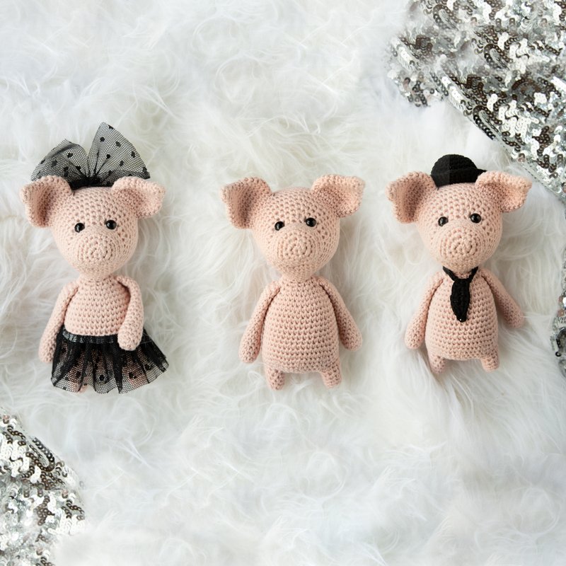 Drei gehäkelte Amigurumi-Schweinchen auf weißem Fell: links mit Tutu und Schleife, Mitte schlicht, rechts mit Hut und Krawatte