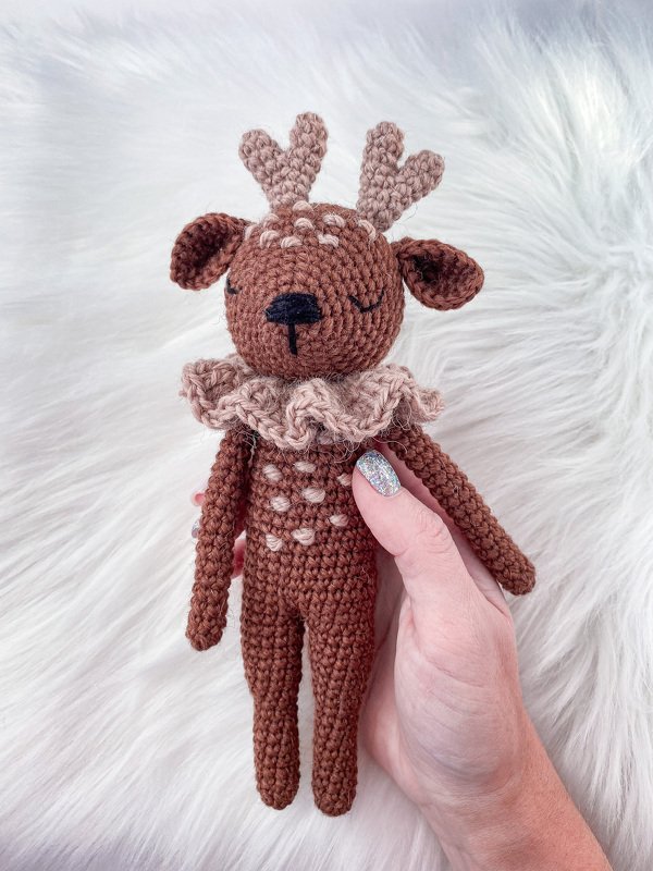 Hand holding small crocheted brown amigurumi deer with antlers and ruffled collar on white faux-fur background
