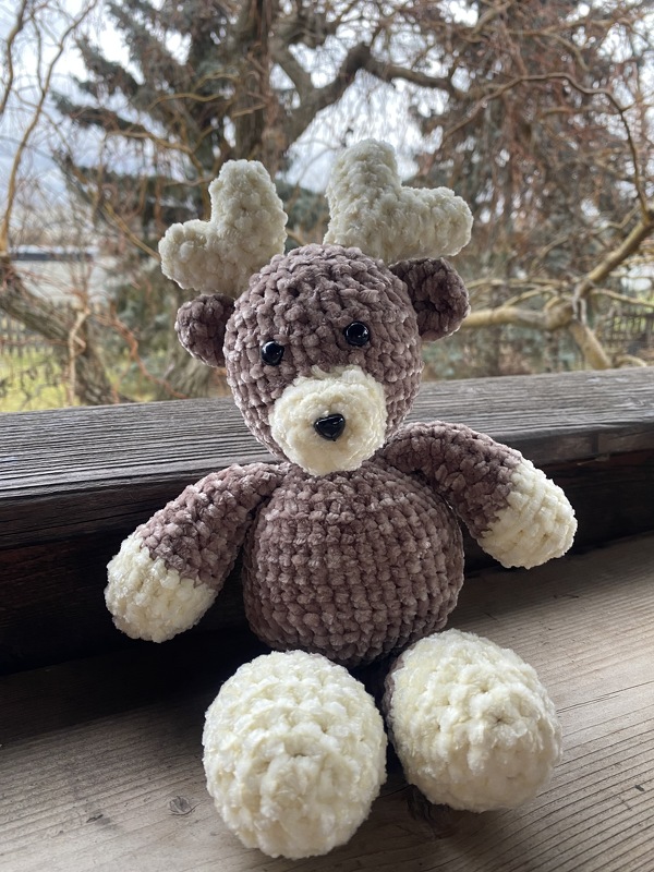 Brown and cream crocheted reindeer plush with cream antlers sitting on a wooden ledge.