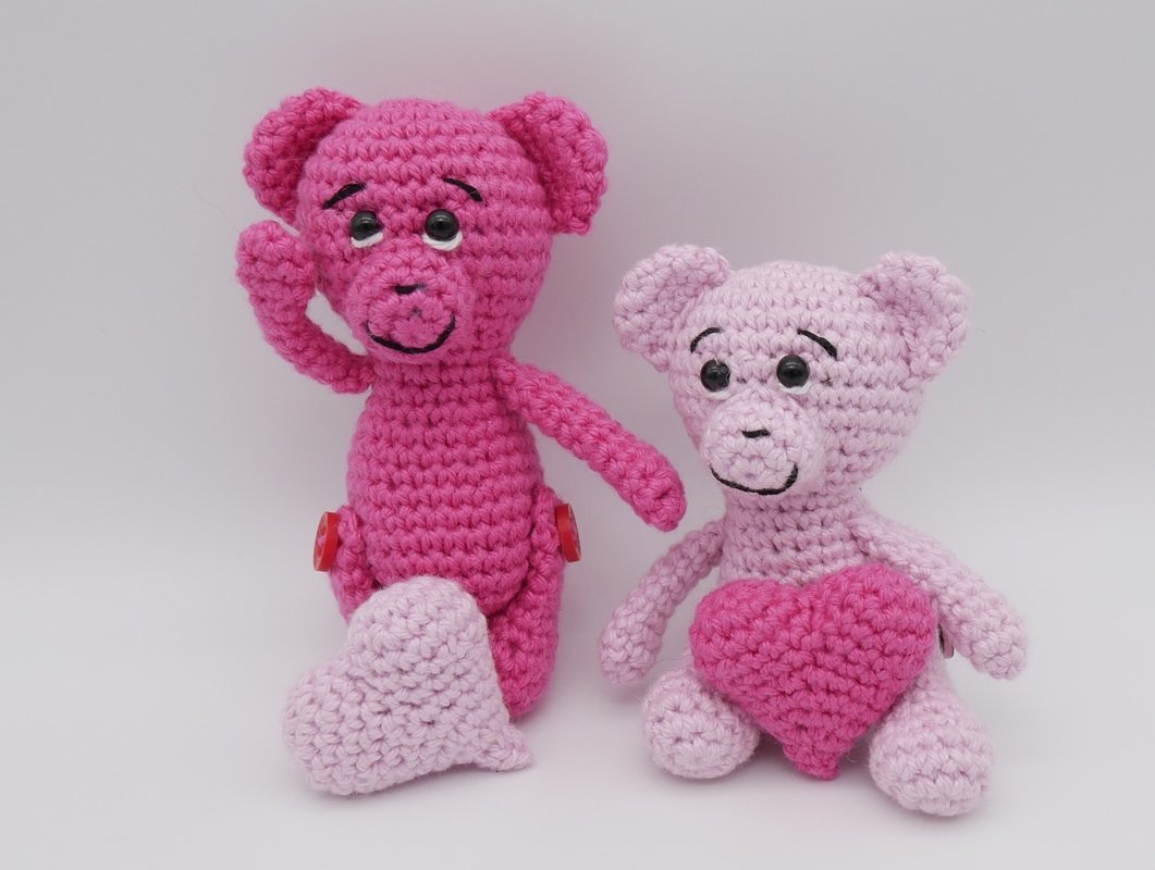 Zwei gehäkelte Amigurumi-Teddys in Pink und Hellrosa, einer mit erhobener Pfote und rotem Knopf an der Hüfte, der andere mit Herz-Applikation auf dem Bauch, vor hellem Hintergrund