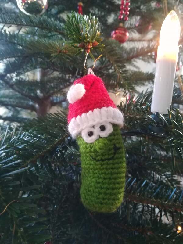 Gehäkelte grüne Gurke mit roter Weihnachtsmütze als Baumschmuck am Tannenzweig.