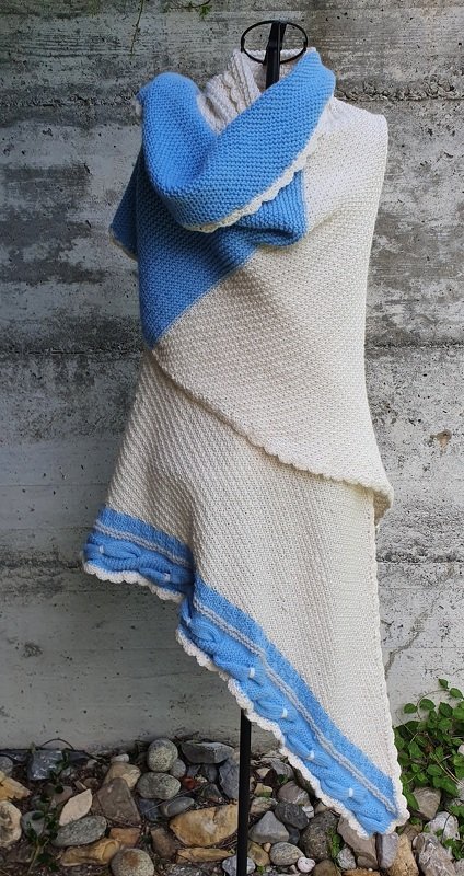 Gestricktes Dreieckstuch in Cremeweiß und Hellblau mit gewelltem Rand, Noppen und Zopfmustern, drapiert an einer Schneiderpuppe vor einer Betonwand