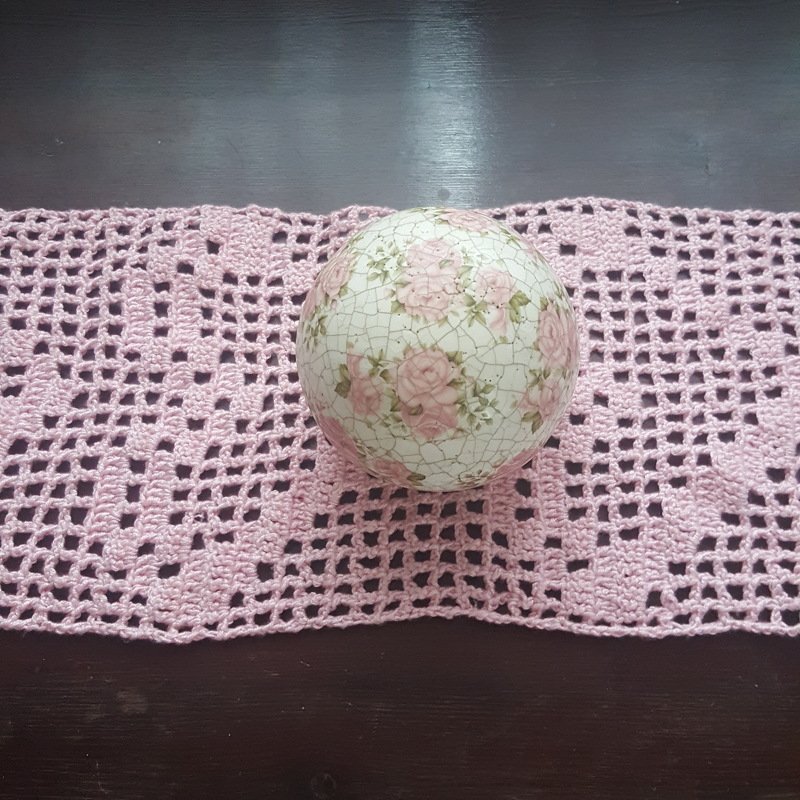 Rosafarbener filetgehäkelter Tischläufer mit wiederholten Herzmotiven und dekorativer Blumenkugel auf dunklem Untergrund