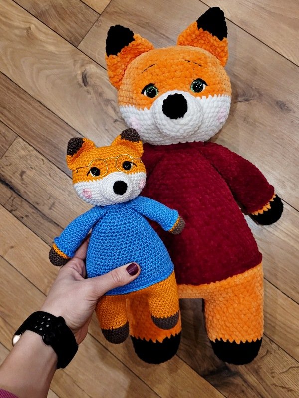 Zwei gehäkelte Fuchsamigurumi – großes mit rotem Pullover, kleines mit blauem Kleid und runder Brille; Hand hält das Kleine