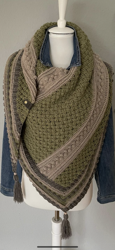 Gehäkeltes Dreieckstuch in Oliv, Beige und Grau mit strukturiertem Muster, Quasten und Verschluss auf einer Schneiderpuppe.