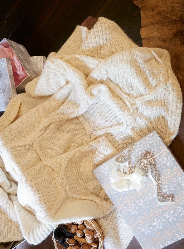 Kuschelige cremeweiße Strickdecke mit Zopfmuster neben dekorativen Geschenkboxen und einem Korb mit Nüssen