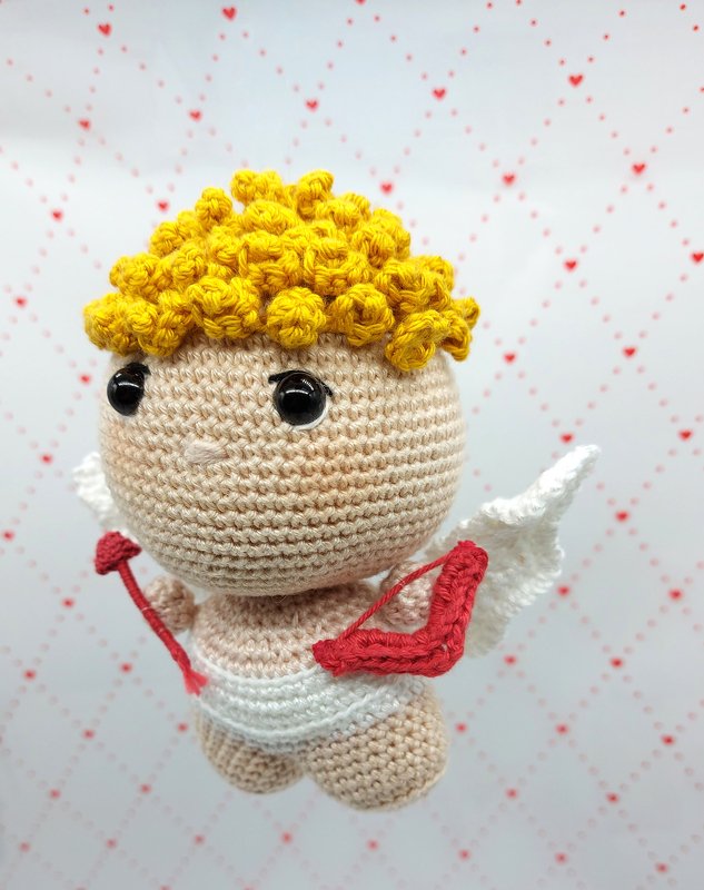 Niedlicher Amigurumi-Amor mit gelbem lockigem Haar, schwarzen Augen, weißen Flügeln und rotem Bogen und Pfeil