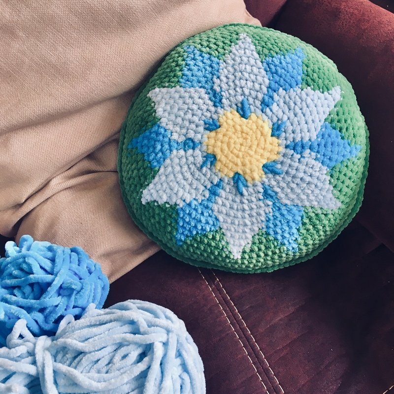 Rundes gehäkeltes Kissen mit blau-weißem Blumenstern auf grünem Hintergrund, hellblaue Wollknäuel daneben, auf Sofa