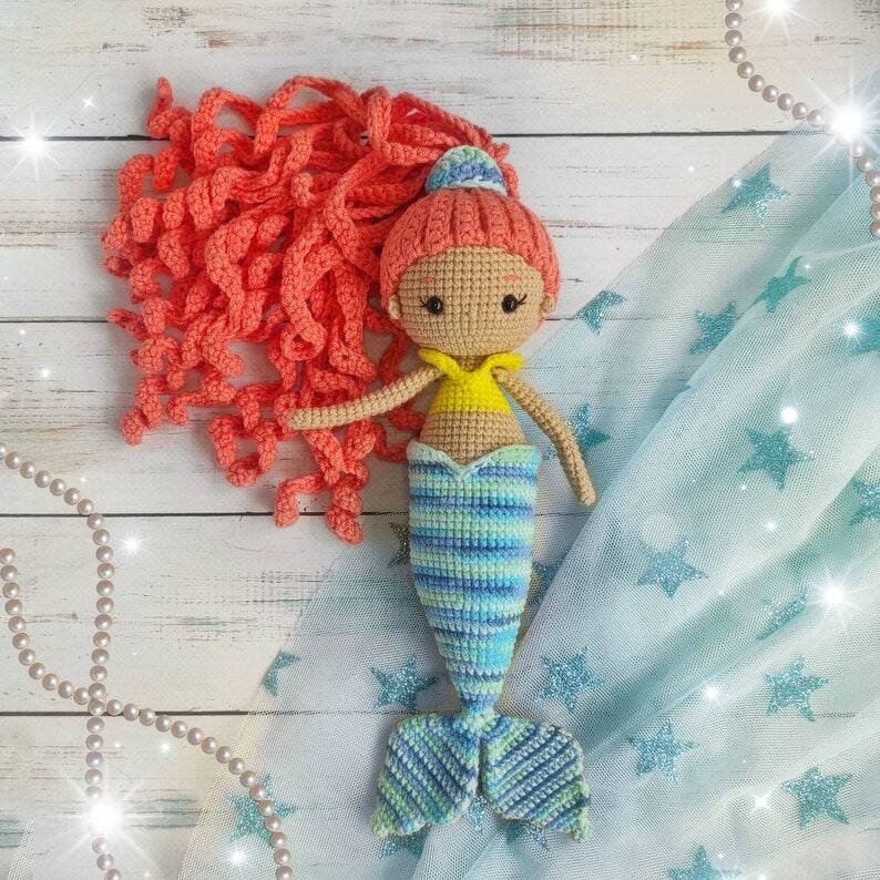 Crocheted amigurumi mermaid doll with coral curly hair and blue-green striped tail on white wood with star tulle