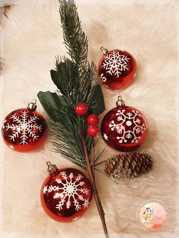 Vier rote Weihnachtskugeln mit weißen Schneeflockenmotiven neben Tannenzweig und Zapfen auf hellem Fell