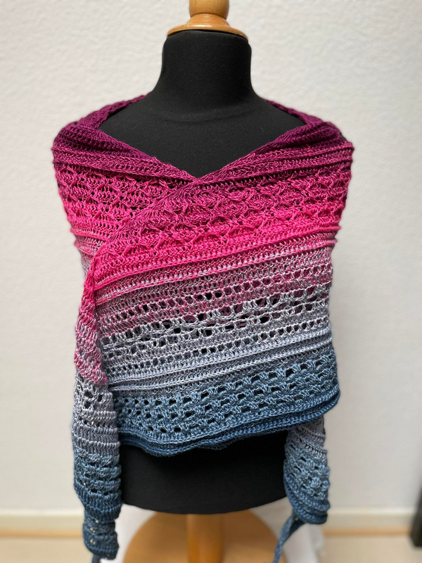 Gehäkeltes Halbrundtuch auf einer Schneiderpuppe mit Farbverlauf von Magenta zu Grau und Blau und verschiedenen Häkelmustern.