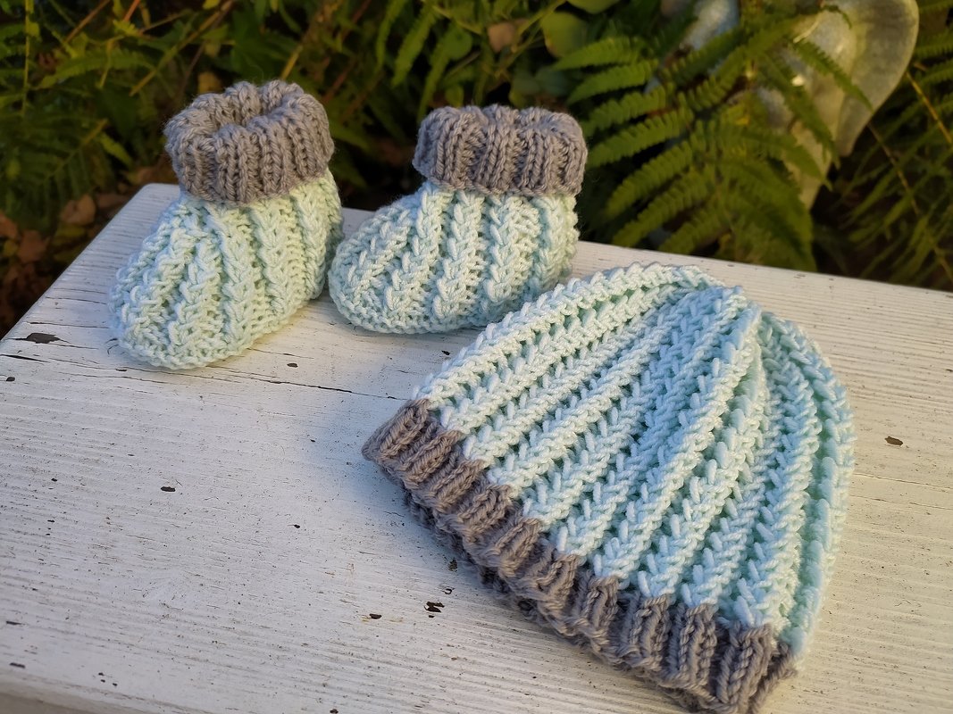 Ein gestricktes Babyset, bestehend aus Mütze und zwei Schühchen in Mintgrün mit grauen Bündchen, liegt auf einem weißen Holzbrett.