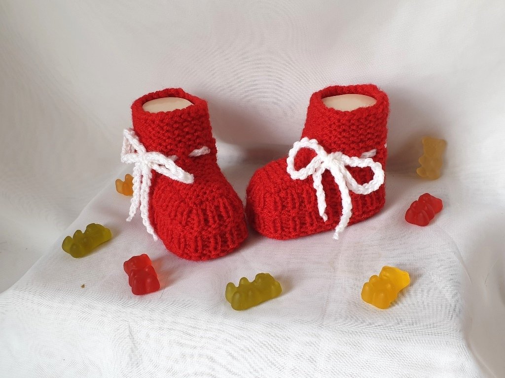 Zwei rote gestrickte Babyschuhe mit weißen Schleifenbändern auf hellem Stoff, dekoriert mit bunten Fruchtgummis
