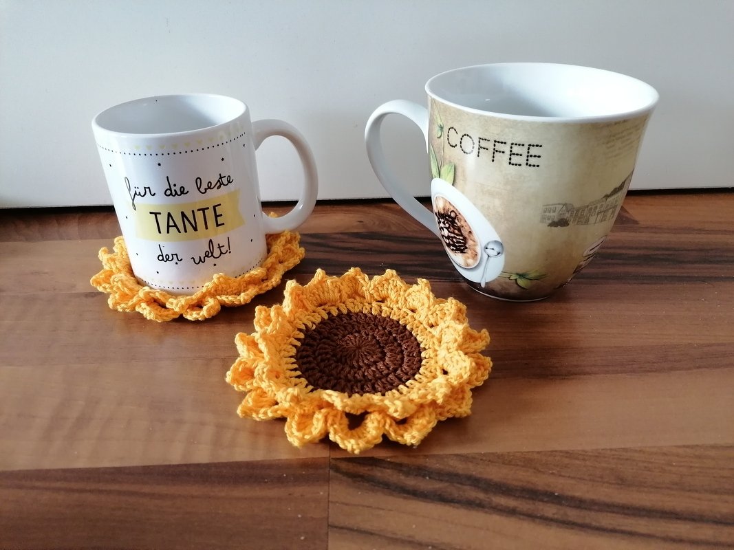 Zwei gehäkelte Untersetzer in Sonnenblumenoptik (braune Mitte, gelbe Rüschen) neben zwei Kaffeetassen auf Holz