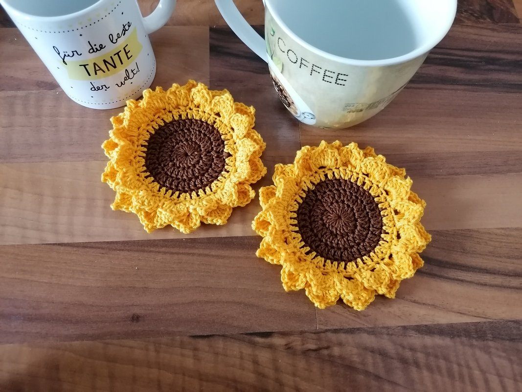 Zwei gehäkelte Sonnenblumen-Untersetzer in Gelb und Braun auf dunklem Holztisch, mit zwei Tassen im Hintergrund.