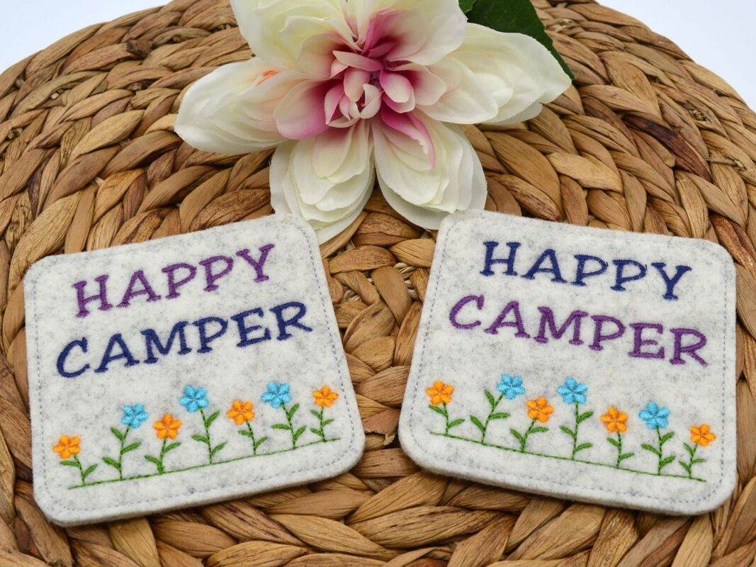 Zwei helle Filz-Tassenteppiche, angefertigt mit der ITH-Stickdatei, zeigen 'HAPPY CAMPER' in lila und blau sowie eine Reihe blauer und oranger Blumen auf einem geflochtenen Naturuntergrund.