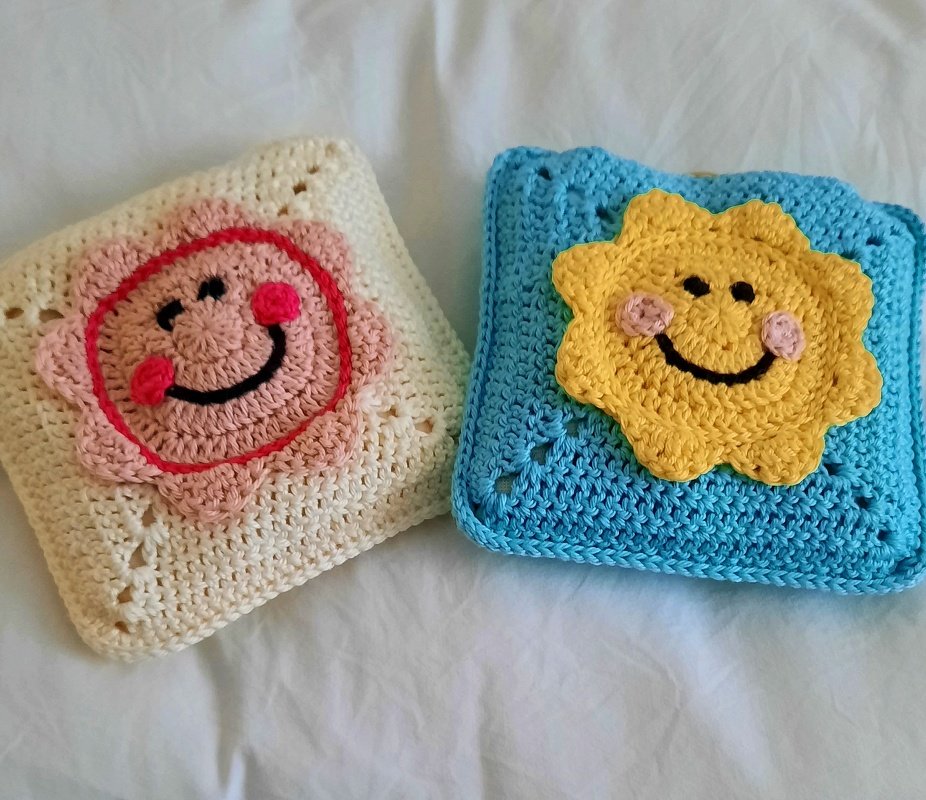 Zwei gehäkelte quadratische Applikationen: cremefarben mit rosa Smiley und hellblau mit gelber Sonnen-Applikation.