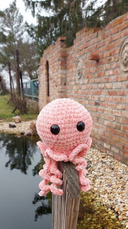 Rosa gehäkelter Tintenfisch-Amigurumi mit großen schwarzen Augen auf einem Holzpfosten vor Teich und Backsteinmauer.