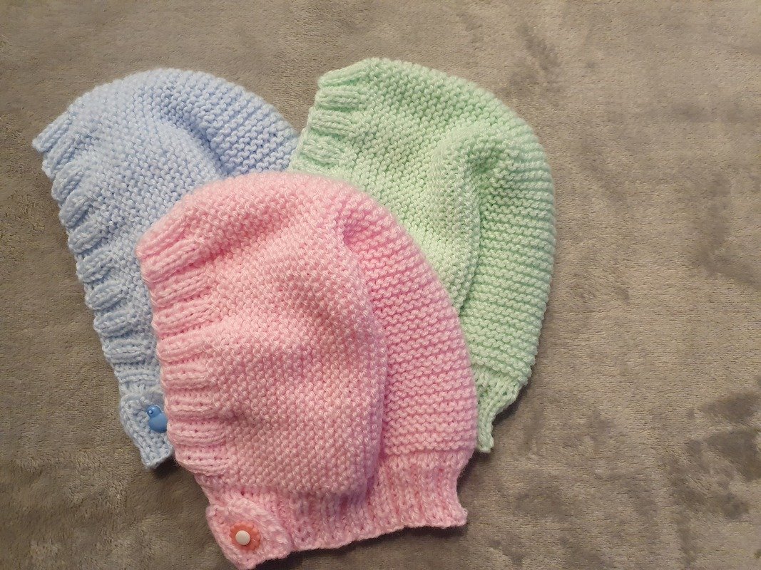 Drei handgestrickte Babymützen in Rosa, Hellblau und Mint mit kleinen Knöpfen auf grauem Stoff