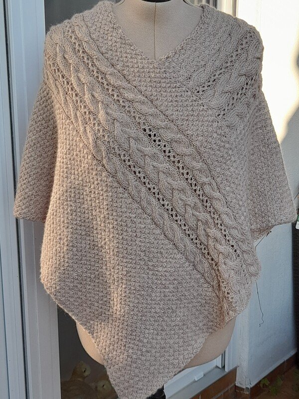 Gestrickter hellbeiger Poncho mit V-Ausschnitt, diagonalen Zopf- und Ajourmustern auf einer Schneiderpuppe.