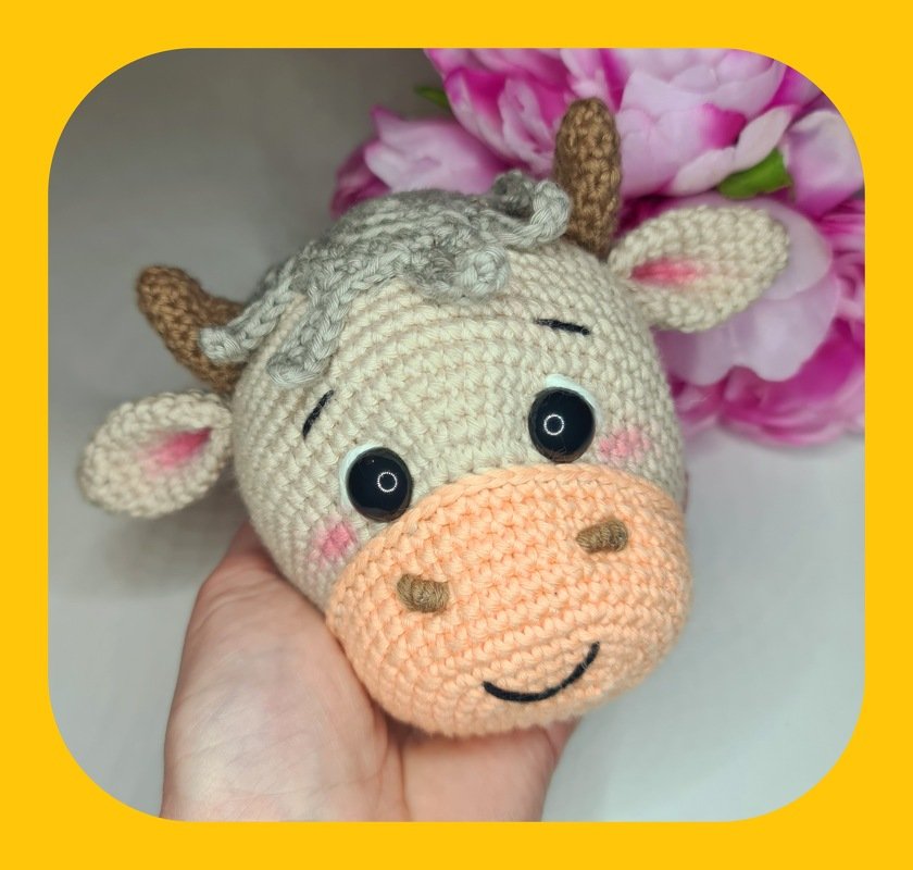 Hand hält gehäkelten Amigurumi-Kuhkopf in Pastellfarben mit schwarzen Augen und rosa Wangen