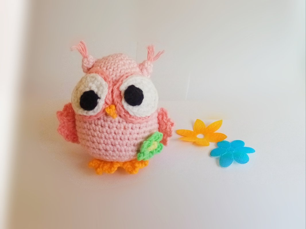 Rosa gehäkelte Amigurumi-Eule mit großen weißen Augen, orangen Füßchen und grüner Blattapplikation, neben bunten Dekoblüten