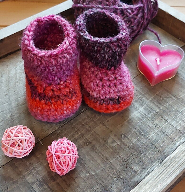 Zwei gehäkelte Babyschuhe aus pink-lila-orangefarbenem Garn auf Holztablett mit passendem Garn und Deko