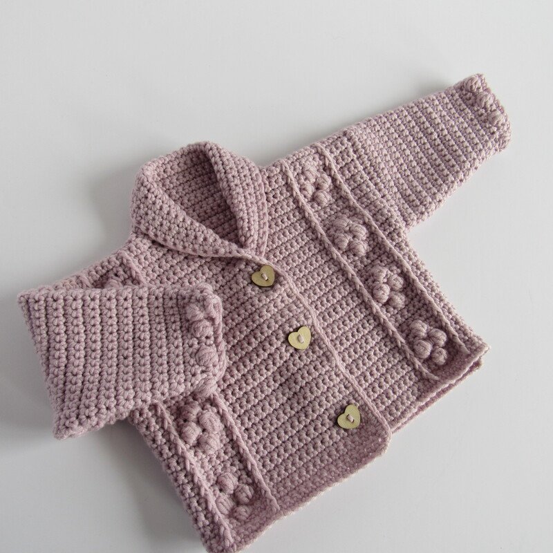 Dusty pink crocheted baby jacket with shawl collar and three heart-shaped buttons, flat-lay