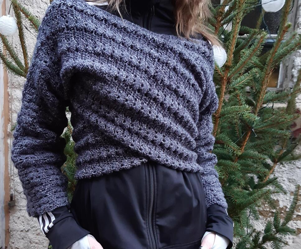 Person trägt dunkelgrauen gehäkelten Schulterwickel mit diagonalem Muster vor Tannenzweigen mit Lichterkette.