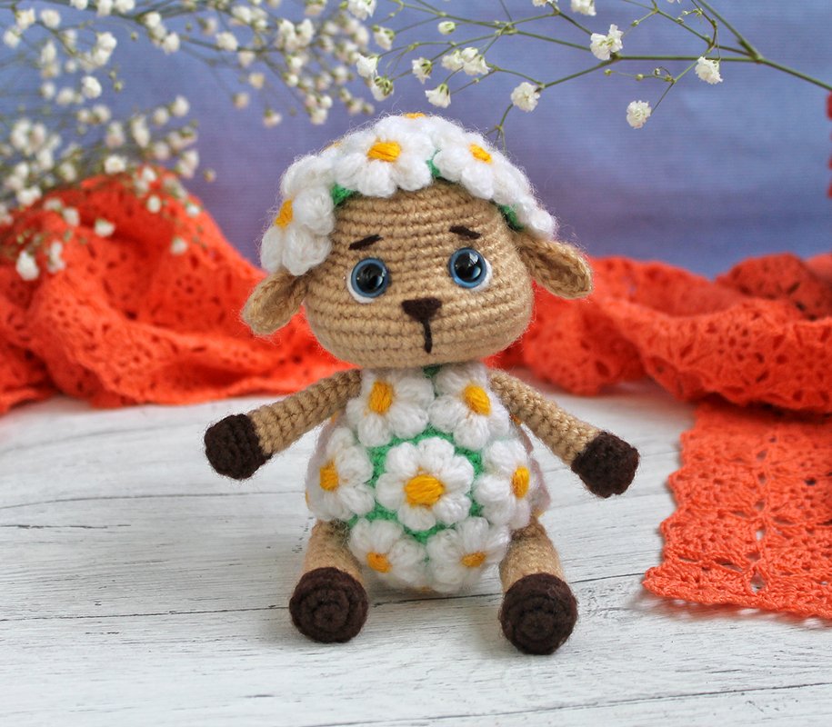 Gehäkeltes Amigurumi‑Schaf mit Gänseblümchen‑Muster, blauen Augen und braunen Hufen auf hellem Holzhintergrund mit orangefarbenem Stoff und weißen Blüten.