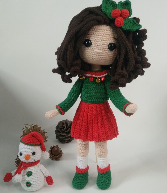 Crocheted amigurumi doll with curly brown hair, green sweater, red pleated skirt, holly hairpiece, bell-trimmed collar, and a small crocheted snowman