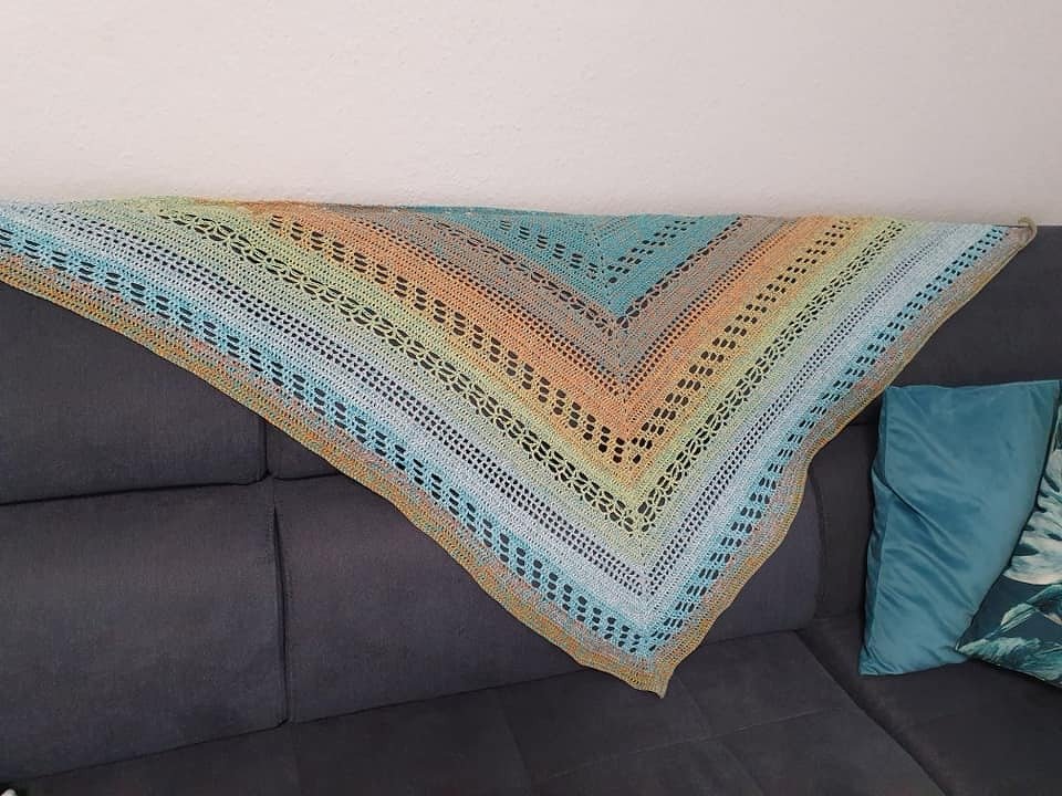 Handgehäkeltes Dreieckstuch mit Farbverlauf und Lochmuster auf grauem Sofa.