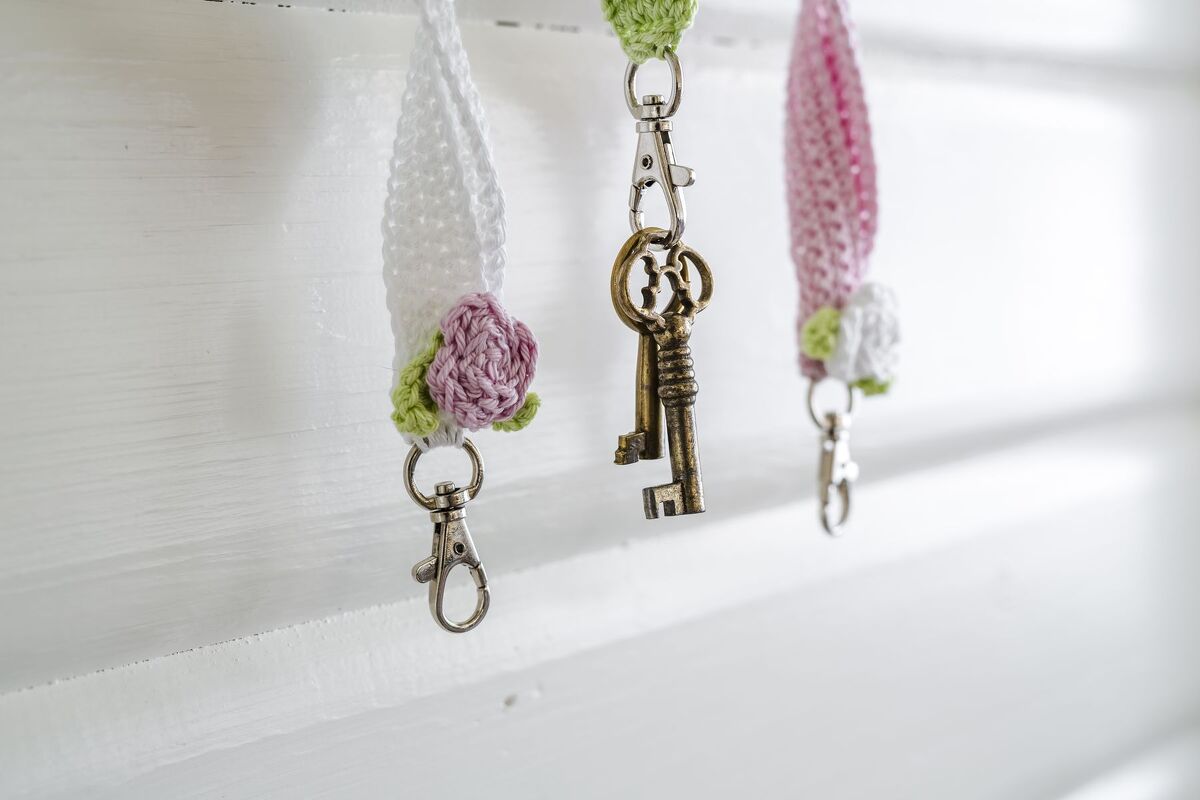 Drei gehäkelte Schlüsselanhänger in Weiß, Grün und Rosa mit Blumenapplikationen; einer trägt zwei dekorative Schlüssel.