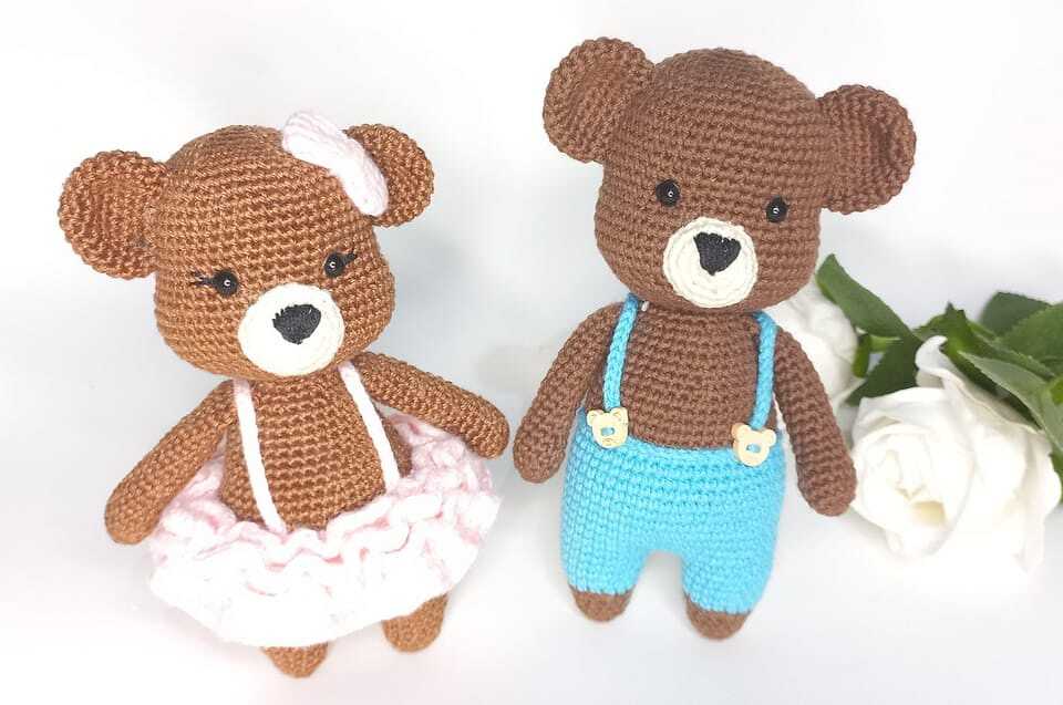 Two brown crocheted amigurumi teddy bears, one in a pink ruffled skirt with a bow and one in blue suspender pants