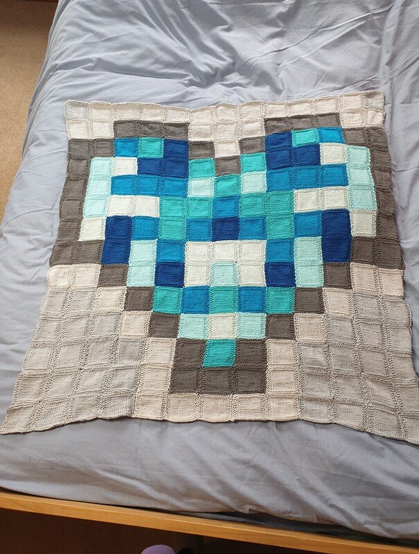 Gestrickte Decke mit großem Pixel-Herz in Blau-, Türkis-, Weiß- und Grautönen auf einem Bett