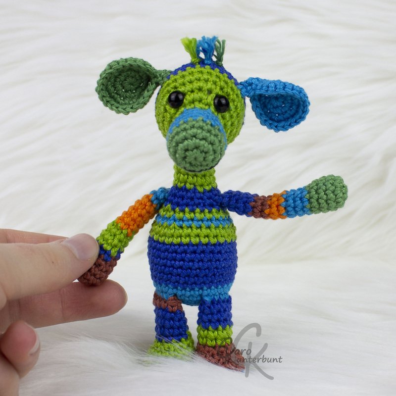 Hand hält kleines, bunt gestreiftes Amigurumi mit grünem Kopf und blauen Ohren auf weißem, flauschigem Hintergrund.