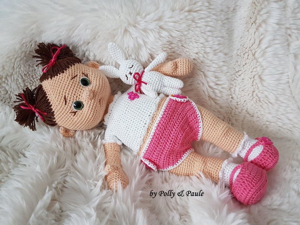 Gehäkelte Puppe mit Zöpfen, weißem Oberteil, rosa Höschen und kleinen rosa Schuhen, liegend auf hellem Fell, hält ein weißes Häschen
