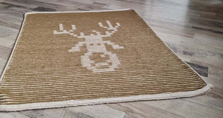 Beige gestrickte Babydecke mit weißem Rentierkopf und heller Kante auf Holzfußboden