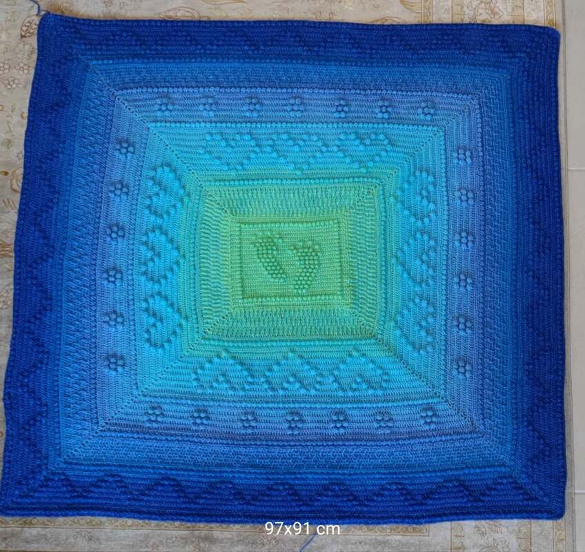 Quadratische gehäkelte Babydecke mit Ombré‑Farbverlauf und zentralen Babyfußabdrücken in Blau‑ und Grüntönen.