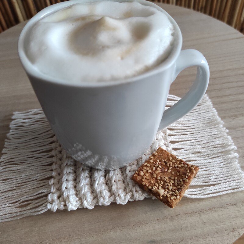 Weiße Tasse mit Milchschaum auf rechteckigem gehäkeltem Untersetzer mit Fransen, daneben Sesamkeks