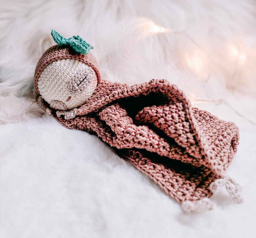 Gehäkelte Babypuppe mit Blattmützchen und gestickten Augen, eingekuschelt in ein rosafarbenes Schmusetuch auf flauschigem Teppich.