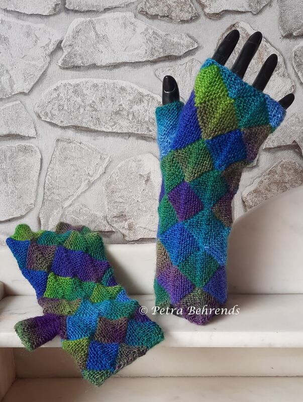 Bunte gestrickte fingerlose Damen-Armstulpen im Entrelac-Rautenmuster auf schwarzer Mannequin-Hand vor hellem Steinmauer-Hintergrund