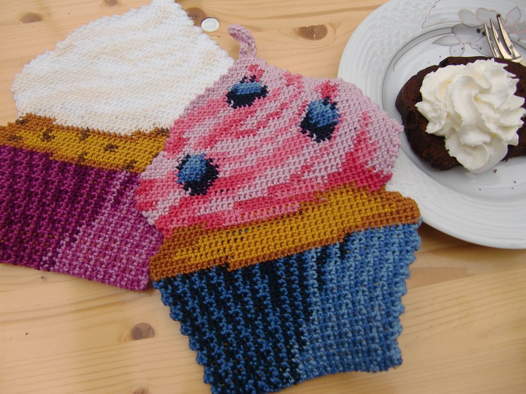 Zwei gehäkelte Topflappen in Cupcake-Form auf einem Holztisch neben einem Teller mit Dessert.